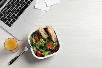 Photo of Container of tasty food, laptop, fork and glass of juice on white wooden table, flat lay with space for text. Business lunch