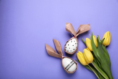 Photo of Easter bunnies made of craft paper and eggs near beautiful tulips on purple background, flat lay. Space for text