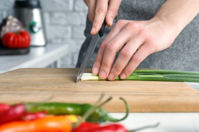 Woman cutting green spring onion on wooden board at white table, closeup