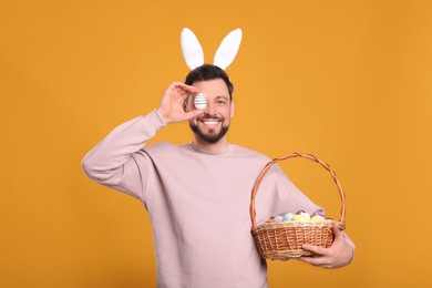 Photo of Happy man in bunny ears headband holding wicker basket with painted Easter eggs on orange background