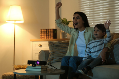 Emotional young woman and her son watching TV using video projector at home. Space for text