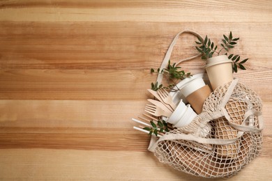 Net bag with disposable tableware and green twigs on wooden background, top view. Space for text