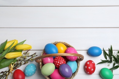 Flat lay composition with colorful Easter eggs in wicker basket on white wooden table. Space for text