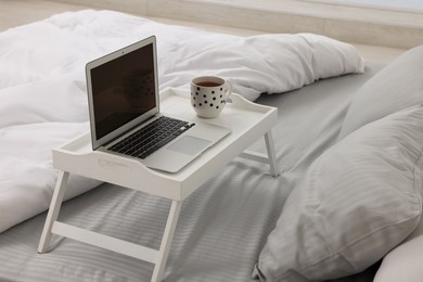White tray table with laptop and cup of drink on bed