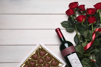 Photo of Bottle of red wine, beautiful roses and heart shaped chocolate candies on white wooden table, flat lay. Space for text