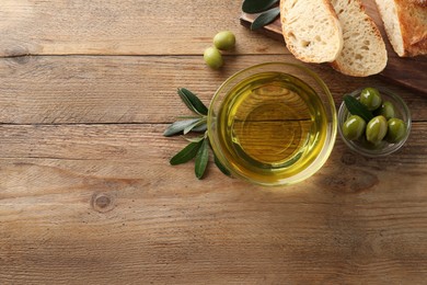 Glass bowl of oil, ripe olives and bread on wooden table, flat lay. Space for text