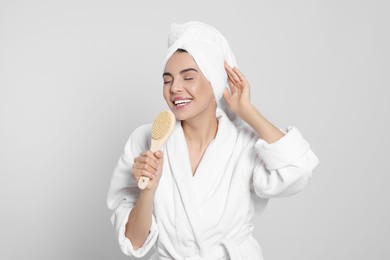 Photo of Woman in bathrobe with towel singing into brush on light grey background. Spa treatment