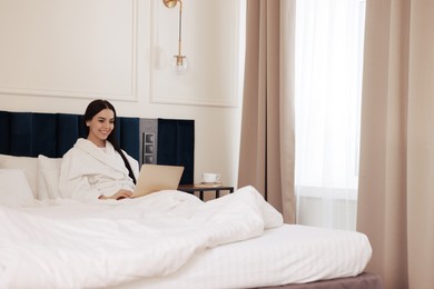 Happy young woman working with laptop on bed in hotel room
