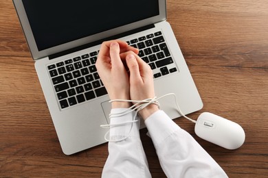 Woman showing hands tied with computer mouse cable near laptop at wooden table, closeup. Internet addiction