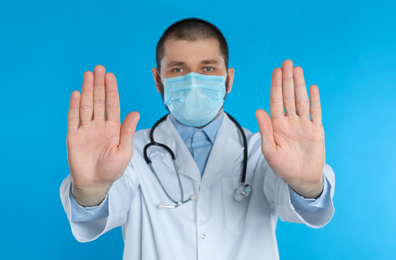 Doctor in protective mask showing stop gesture on light blue background. Prevent spreading of coronavirus