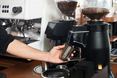 Barista with metal cup using coffee grinding machine in cafe, closeup