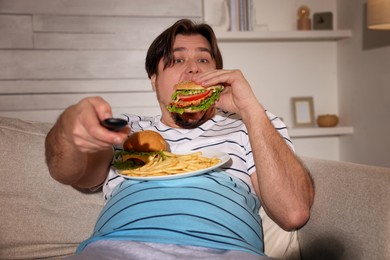 Overweight man with plate of burgers and French fries watching TV on sofa at home