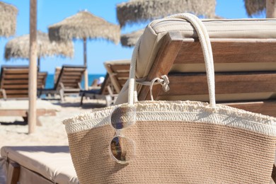 Straw bag with sunglasses on wooden sunbed near sea, closeup. Beach accessories