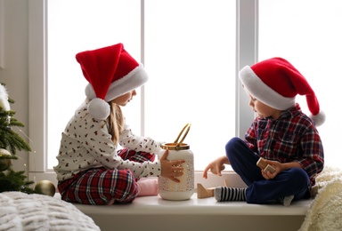 Photo of Cute little children in Santa hats playing on window sill near Christmas tree at home