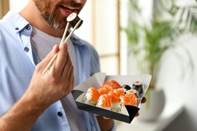 Handsome man eating sushi rolls with chopsticks indoors, closeup
