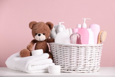 Baby cosmetic products in wicker basket, bath accessories and knitted toy bear on white table against pink background