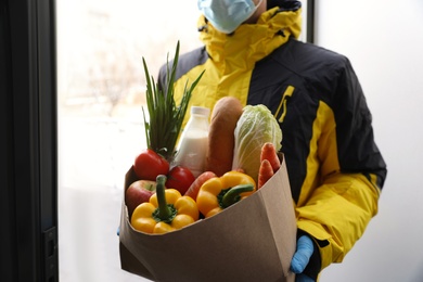 Courier in medical mask holding paper bag with groceries at doorway, closeup. Delivery service during quarantine due to Covid-19 outbreak