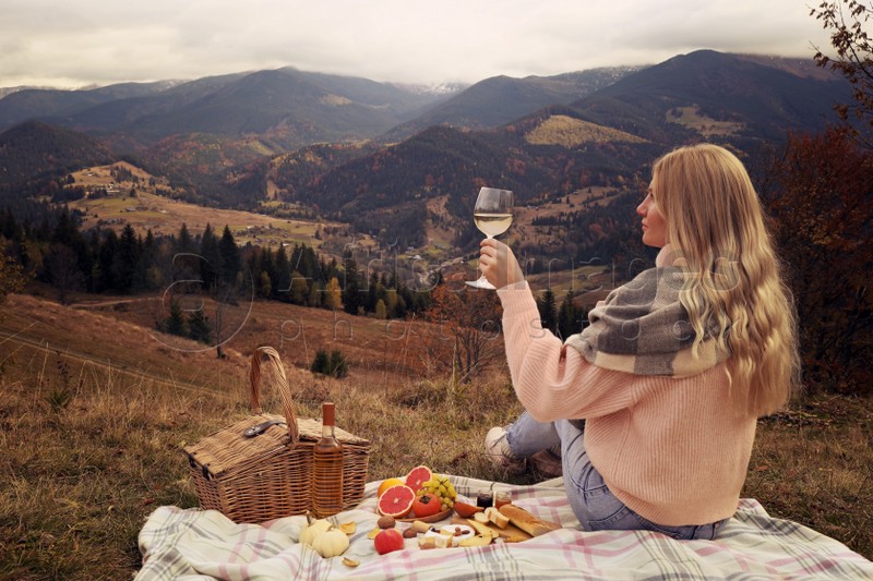Young woman with glass of wine having picnic in mountains on autumn day