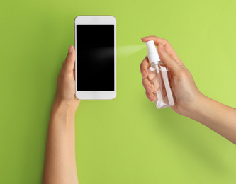 Woman sanitizing smartphone with antiseptic spray on green background, closeup. Be safety during coronavirus outbreak 