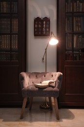 Comfortable armchair with book and lamp between wooden bookcases in library
