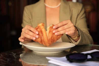 Woman breaking tasty croissant at table in cafeteria, closeup