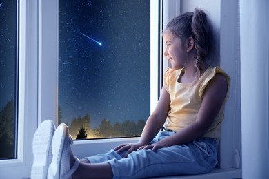 Cute little girl sitting near window and looking at shooting star in beautiful night sky