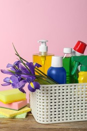 Photo of Spring cleaning. Basket with detergents, flowers and sponges on wooden table against pink background