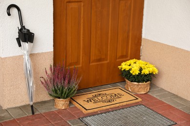 Door mat with word Welcome, umbrella and beautiful flowers near entrance