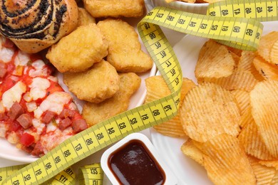 Photo of Different unhealthy food and measuring tape on white table, top view. Weight loss concept