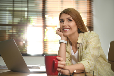 Young woman with cup of drink relaxing at workplace