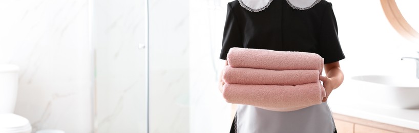 Young chambermaid holding stack of fresh towels in bathroom, closeup view with space for text. Banner design