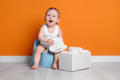 Photo of Little child sitting on baby potty and box of diapers near orange wall