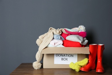 Donation box with knitted clothes and rubber boots on table near grey wall. Space for text