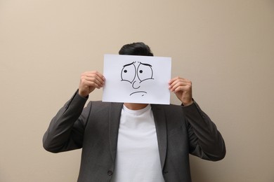 Man hiding emotions using card with drawn frowning face on beige background