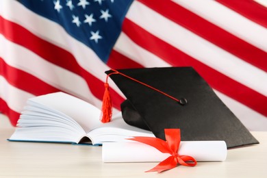 Photo of Black graduation hat, diploma and book on white wooden table against American flag