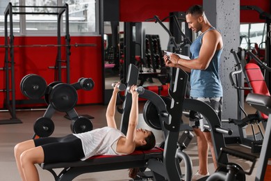Photo of Trainer writing down plan of workouts while woman doing exercise in modern gym