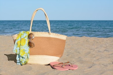 Straw bag with beach wrap, sunglasses and flip flops on sandy seashore, space for text. Summer accessories