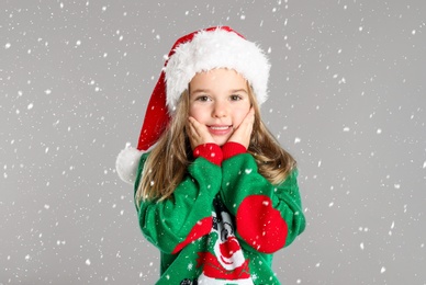 Cute child in Santa hat under snowfall on grey background. Christmas celebration