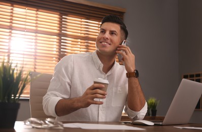 Freelancer with cup of coffee talking on phone while working at table indoors