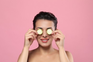 Woman covering eyes with cucumber slices on pink background. Skin care