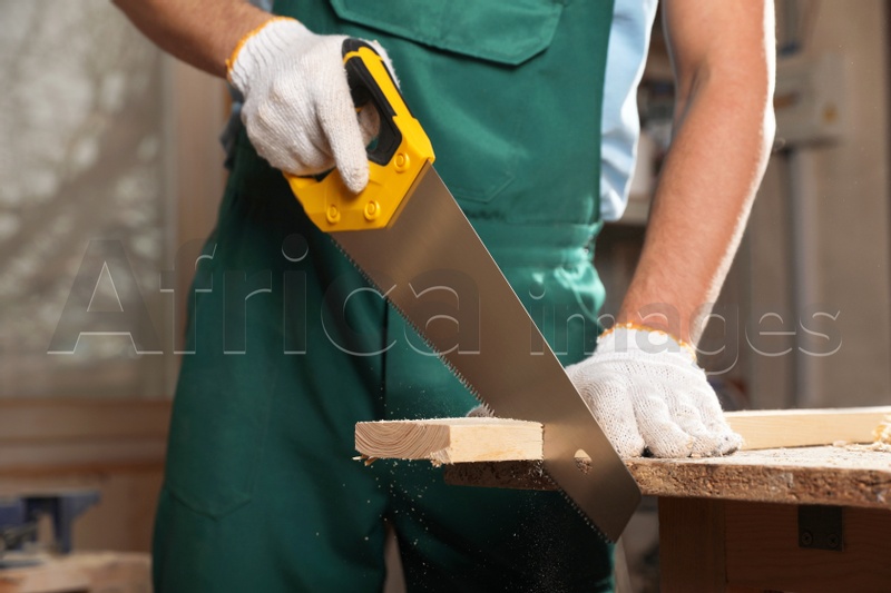Professional carpenter cutting wooden board with handsaw in workshop, closeup