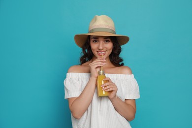 Beautiful young woman with straw hat and bottle of refreshing drink on light blue background