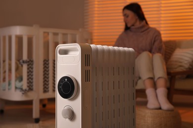 Photo of Young woman near crib and modern portable electric heater indoors, focus on device