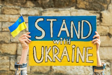 Woman holding poster in colors of national flag and words Stand With Ukraine against brick wall outdoors, closeup