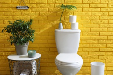 Photo of Stylish bathroom with toilet bowl, green plants and decor elements near yellow brick wall. Interior design