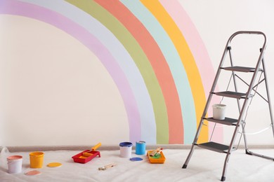Different decorator's tools and ladder near wall with painted rainbow indoors