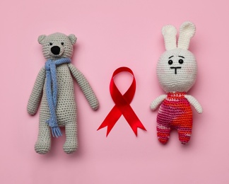Photo of Cute knitted toys and red ribbon on pink background, flat lay. AIDS disease awareness