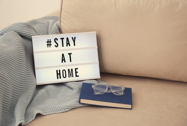 Book, glasses, plaid and lightbox with hashtag STAY AT HOME on sofa. Message to promote self-isolation during COVID‑19 pandemic