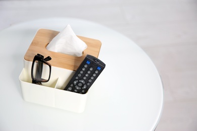 Box with paper tissues, glasses and TV remote control on white table. Space for text