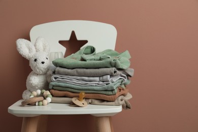 Photo of Stack of baby clothes, pacifier and toys on chair near light brown wall. Space for text